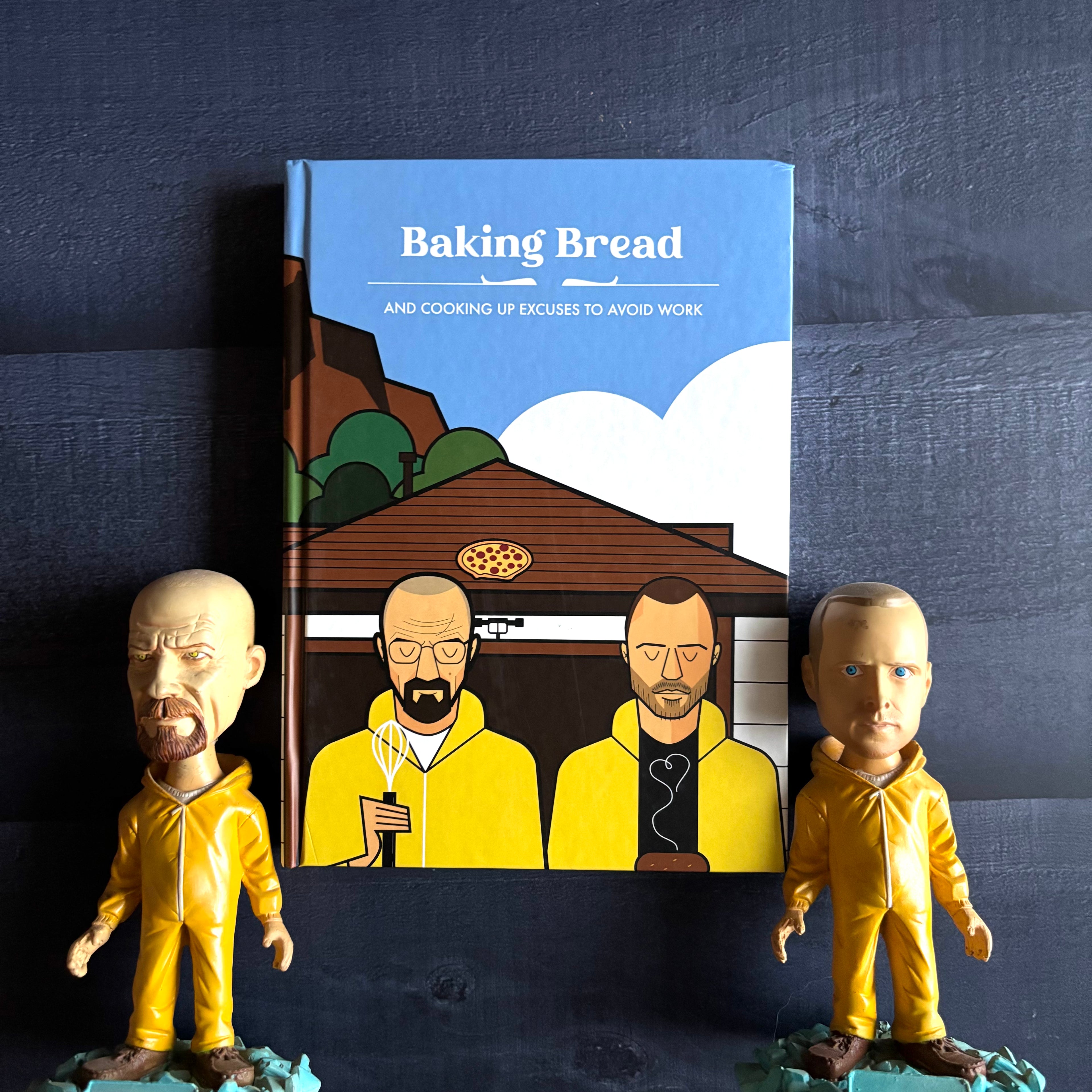 Book titled 'Baking Bread' with illustrations of Walter White and Jessie Pinkman in yellow suits, accompanied by bobblehead figures of these characters on a dark blue background.