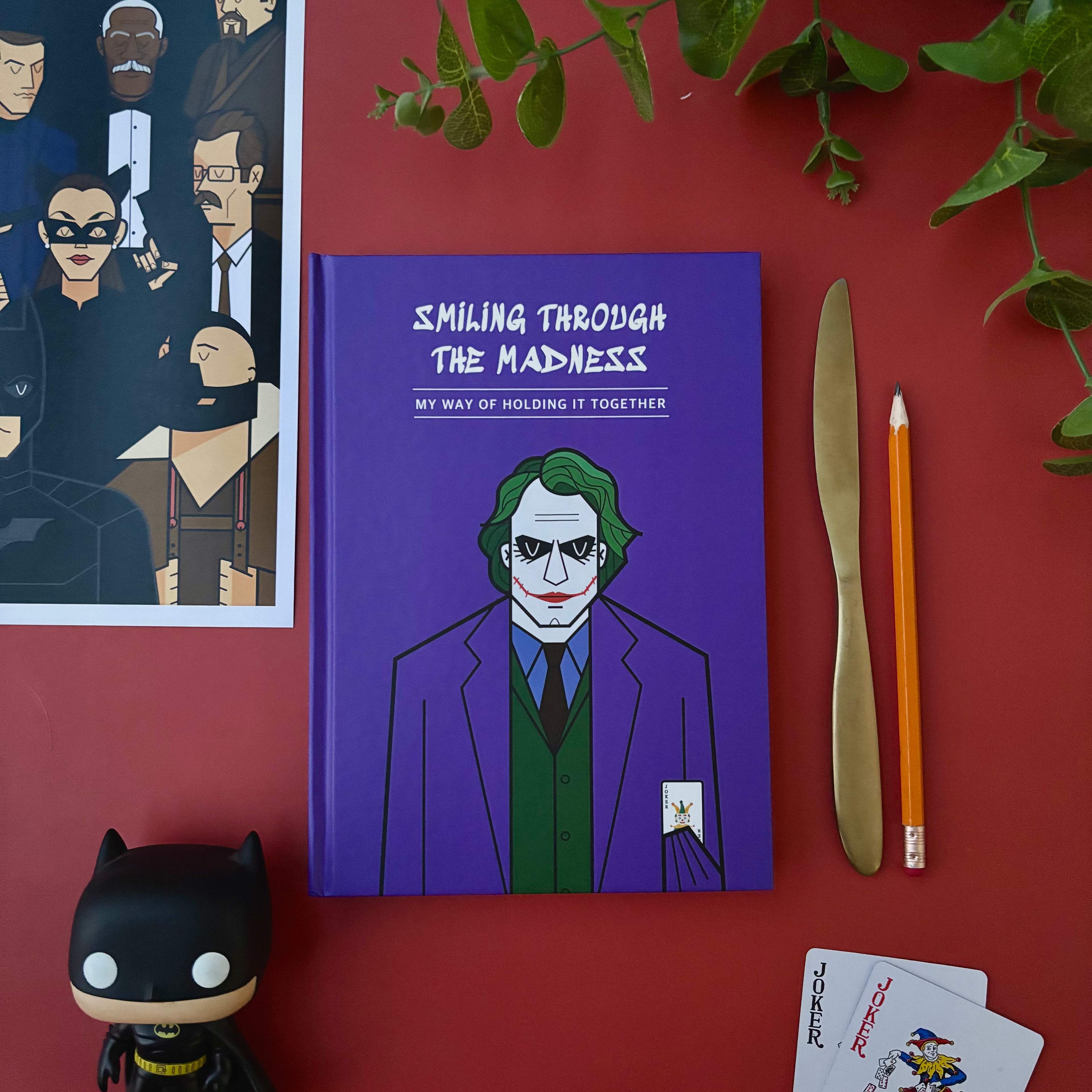 Purple notebook with Joker illustration on a red surface with Batman figurine and playing cards.  Designed by Monkart.