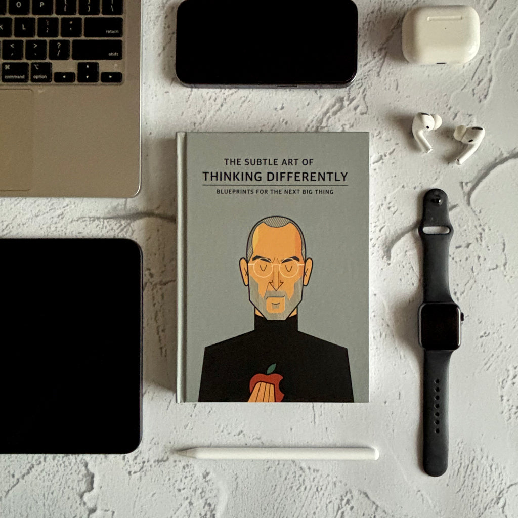 Book titled 'The Subtle Art of Thinking Differently' on a desk with a laptop, phone, and smartwatch. Designed by Monkart.